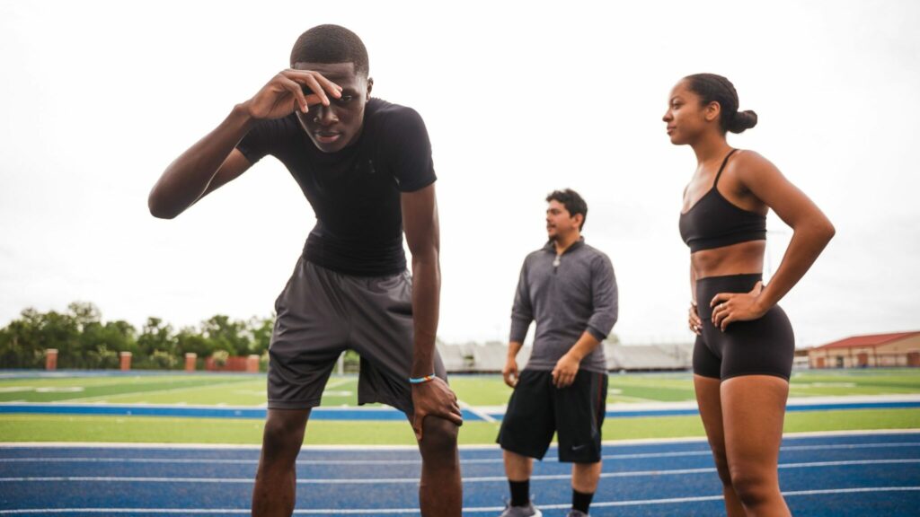 Trois personnes en tenue de sport sur une piste d’athlétisme, dont un homme penché vers l’avant